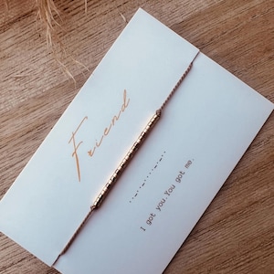 Può includere: Un biglietto bianco con la parola "Friend" scritta in corsivo in alto. Un braccialetto di perline dorate è avvolto attorno al biglietto. Il biglietto contiene anche il testo "I got you. You got me."