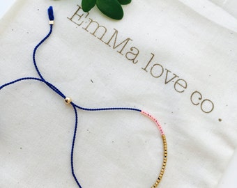 Könnte beinhalten: Ein blaues und goldenes Perlenarmband mit einem rosafarbenen Akzent. Das Armband ist verstellbar und hat eine goldene Perle am Ende der Schnur. Das Armband liegt auf einem weißen Hintergrund mit dem Text "Emma love co."