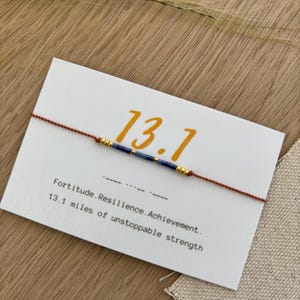 May include: A minimalist bracelet with a brown cord and blue and gold beads, displayed on a white card. The card features the number "13.1" in orange and the words "Fortitude. Resilience. Achievement. 13.1 miles of unstoppable strength."