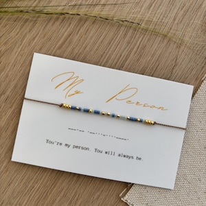 Pode incluir: Um cartão branco com a escrita dourada "My Person" e uma pulseira de fio castanho com contas azuis e douradas. O cartão também tem o texto "You're my person. You will always be."