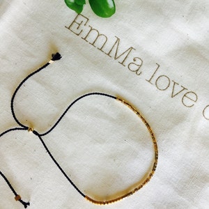 May include: A black cord bracelet with gold beads. The bracelet is adjustable and has a sliding knot. The bracelet is on a white fabric background with the text "Emma Loved" embroidered on it.