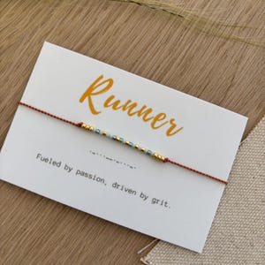 Puede incluir: Una pulsera minimalista con cuentas doradas y turquesas ensartadas en un fino cordón marrón rojizo. La pulsera descansa sobre una tarjeta blanca con la palabra "Runner" en escritura naranja y la frase "Fueled by passion, driven by grit."