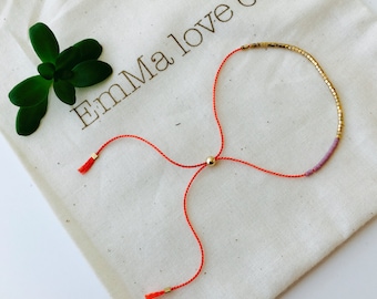 Könnte beinhalten: Ein rotes Armband mit einem goldenen und violetten Perlenanhänger. Das Armband ist mit einem Knoten gebunden und hat an den Enden Quasten. Das Armband liegt auf einem weißen Stoffhintergrund mit dem Text "Emma love co."