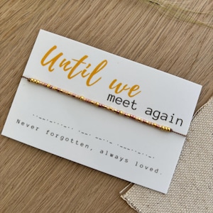 May include: A white card with the text "Until we meet again" in orange script, and "Never forgotten, always loved" in black. A beaded bracelet with gold and pink beads is attached to the card. The card rests on a wooden surface.