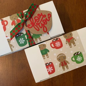 May include: Two rectangular white gift boxes wrapped in a beige paper band featuring a repeating pattern of gingerbread people and mugs of hot cocoa with snowflakes. One box has a red and green plaid ribbon and a brown tag with the text for you.