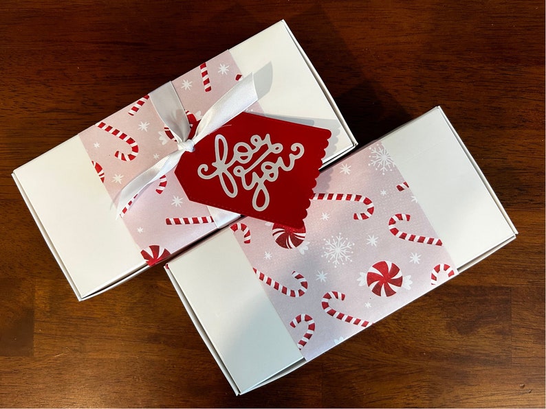 May include: Two rectangular white gift boxes wrapped with a light pink paper band featuring a pattern of red and white candy canes, peppermint swirls, and snowflakes. A white ribbon ties one box, which also has a red tag with the text for you.