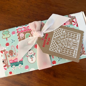 May include: A rectangular white box wrapped in light blue paper featuring a festive pattern of gingerbread houses, snowmen, and candy canes. A pale pink ribbon ties a brown scalloped gift tag with a white gingerbread house and the text a sweet treat for you.