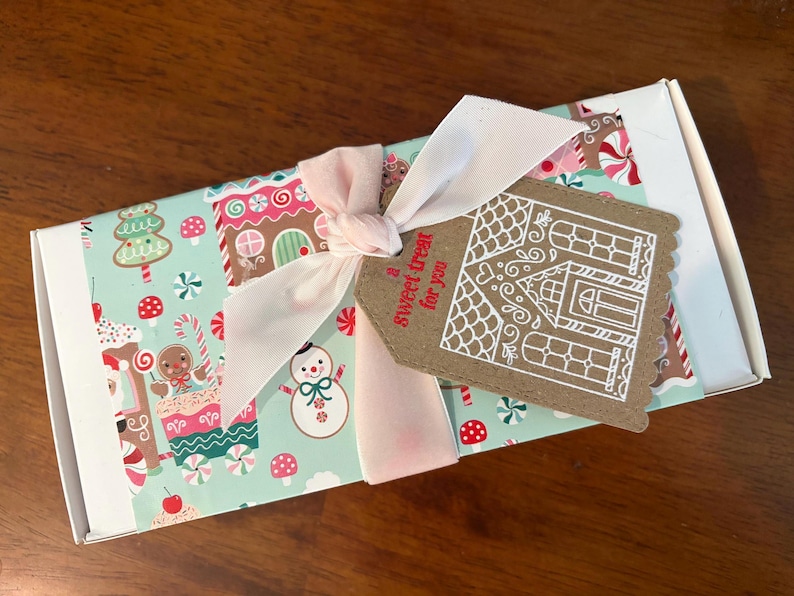 May include: A rectangular white box wrapped in light blue paper featuring a festive pattern of gingerbread houses, snowmen, and candy canes. A pale pink ribbon ties a brown scalloped gift tag with a white gingerbread house and the text a sweet treat for you.
