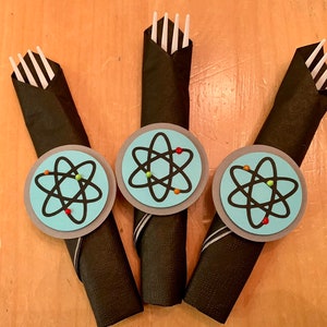 May include: Three black napkins with white forks wrapped inside. Each napkin has a blue circle with a black atom symbol and colorful dots around it.