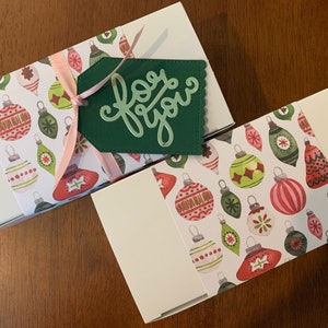 May include: Two rectangular white gift boxes wrapped with a paper band featuring a colorful pattern of Christmas ornaments in red, green, and white. A light pink ribbon ties a dark green scalloped gift tag with the words for you in cursive script.
