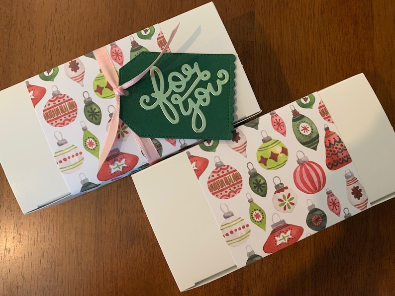 May include: Two rectangular white gift boxes wrapped with a paper band featuring a colorful pattern of Christmas ornaments in red, green, and white. A light pink ribbon ties a dark green scalloped gift tag with the words for you in cursive script.