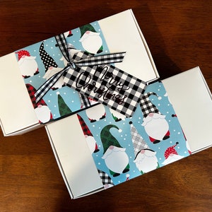 May include: Two rectangular white gift boxes wrapped with a light blue paper band featuring a pattern of gnomes in various hats. A black and white buffalo plaid ribbon ties a matching gift tag with the text Merry Christmas written in black script.