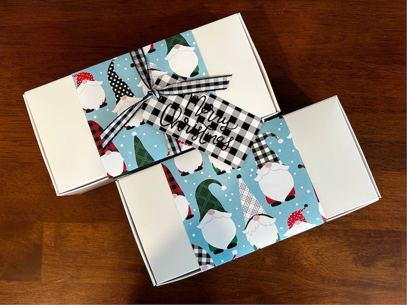 May include: Two rectangular white gift boxes wrapped with a light blue paper band featuring a pattern of gnomes in various hats. A black and white buffalo plaid ribbon ties a matching gift tag with the text Merry Christmas written in black script.