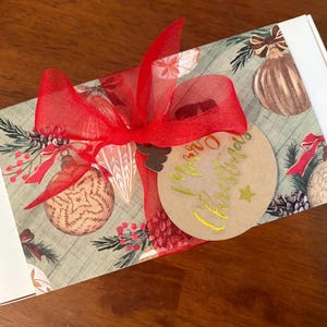 May include: A rectangular white gift box wrapped in a paper sleeve featuring a pattern of Christmas ornaments, pine branches, and berries. A bright red sheer ribbon is tied in a bow, holding a round tan gift tag with gold Merry Christmas text.