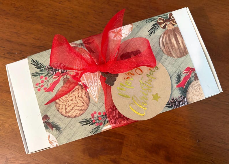 May include: A rectangular white gift box wrapped in a paper sleeve featuring a pattern of Christmas ornaments, pine branches, and berries. A bright red sheer ribbon is tied in a bow, holding a round tan gift tag with gold Merry Christmas text.