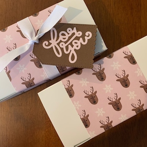 May include: Two rectangular white boxes wrapped in a light pink paper band featuring a repeating pattern of brown reindeer heads and white snowflakes. One box is tied with a white ribbon and a brown scalloped tag with the text for you.