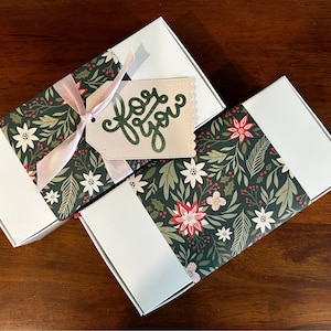 May include: Two rectangular white gift boxes wrapped with a dark green paper band featuring a pattern of white and red poinsettias, holly leaves, and berries. One box has a light pink ribbon and a beige tag with the text for you in green.