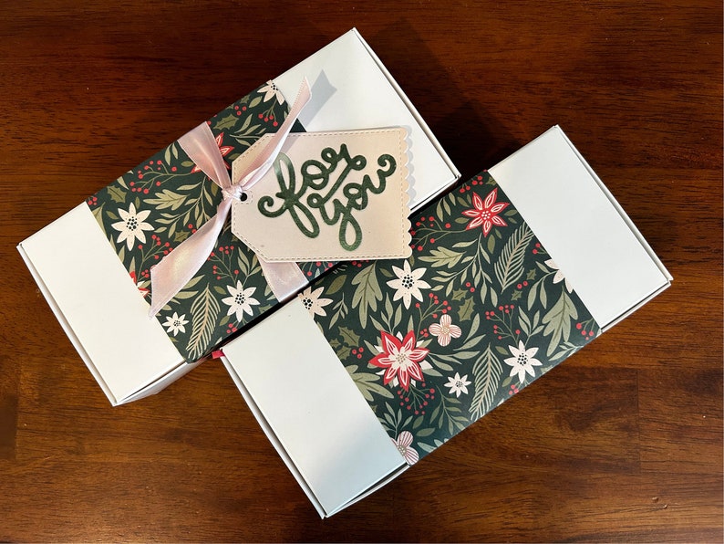 May include: Two rectangular white gift boxes wrapped with a dark green paper band featuring a pattern of white and red poinsettias, holly leaves, and berries. One box has a light pink ribbon and a beige tag with the text for you in green.
