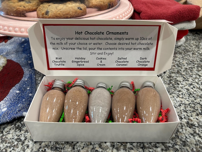 May include: A white gift box containing five clear, lightbulb-shaped plastic ornaments filled with hot chocolate mix. The lid features printed instructions and flavor labels: Mint Chocolate Truffle, Holiday Gingerbread Spice, Cookies & Cream, Salted Chocolate Caramel, and Dark Chocolate Orange. Red and green paper shreds surround the ornaments.
