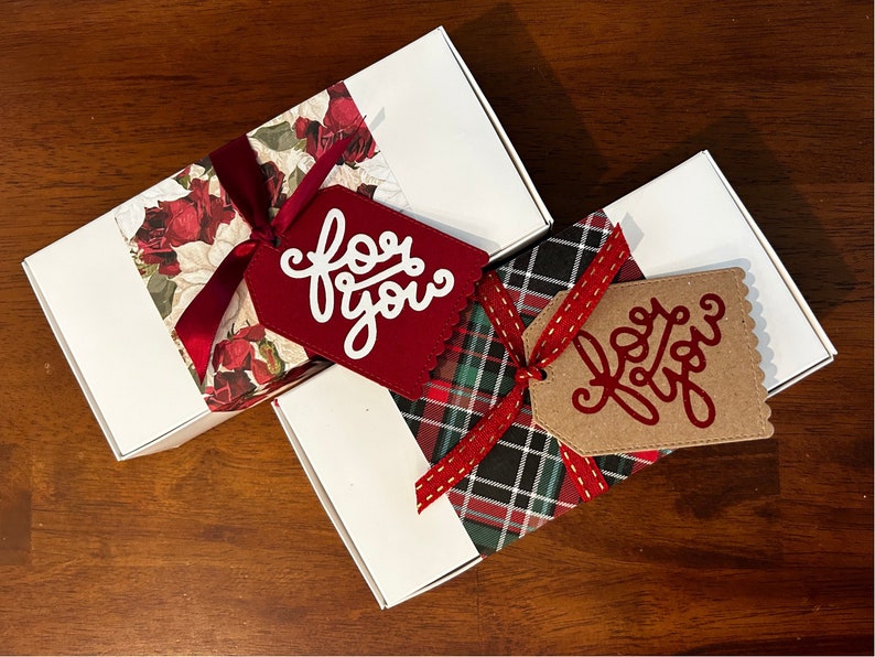 May include: Two white rectangular gift boxes. One box features a red rose floral pattern band with a dark red ribbon and a red tag with the white text for you. The other box has a green and red plaid band with a red ribbon and a tan tag with the red text for you.