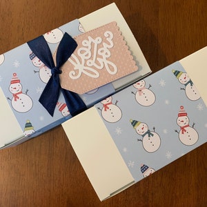 May include: Two rectangular white gift boxes wrapped with light blue paper featuring a repeating pattern of snowmen wearing colorful hats and scarves. The top box includes a dark blue ribbon bow and a scalloped pink tag with the text for you.