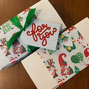 May include: Two white rectangular gift boxes wrapped with a festive paper band featuring a pattern of gnomes, Christmas trees, and presents. A bright green satin ribbon ties one box, accented by a light blue tag with red text reading for you.