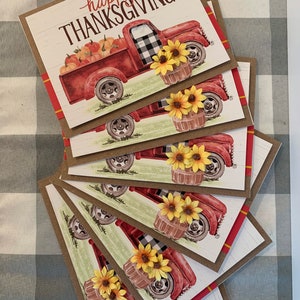 May include: Set of six red and white Thanksgiving cards featuring a vintage red truck with a black and white checkered design, carrying pumpkins and sunflowers. The cards have a rustic brown background with a white woodgrain pattern and the text "happy THANKSGIVING".