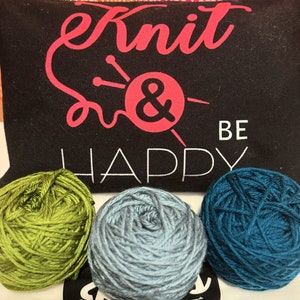 May include: Black zippered pouch with the words "Knit & Be Happy" in pink and white. Three balls of yarn in green, gray, and teal are in front of the pouch. The pouch has a pink ampersand symbol with knitting needles.