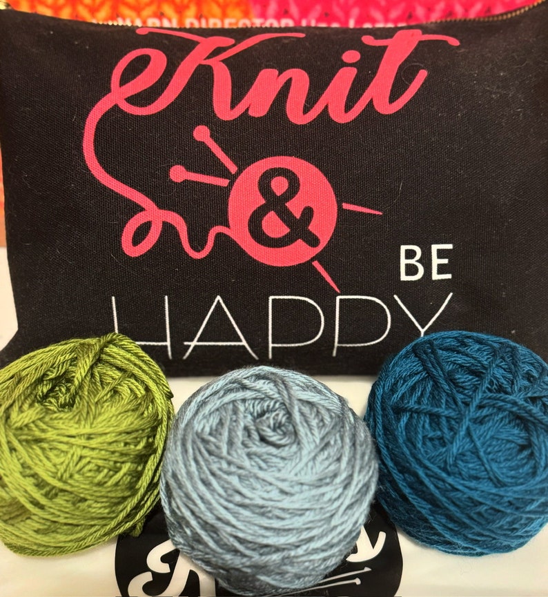 May include: Black zippered pouch with the words "Knit & Be Happy" in pink and white. Three balls of yarn in green, gray, and teal are in front of the pouch. The pouch has a pink ampersand symbol with knitting needles.