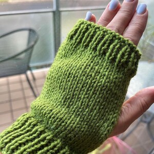 May include: A green knitted fingerless glove, showcasing a ribbed cuff and a textured stitch pattern. The glove is a vibrant shade of green, perfect for keeping hands warm while allowing finger movement. The item is handmade.