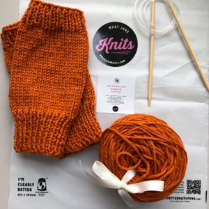 Peut inclure: Un ensemble de chauffe-bras tricotés orange, une pelote de laine assortie, des aiguilles à tricoter et un autocollant indiquant "What Jane Knits". Une petite carte indique "wjk aussie yarn beginner knit kits". Le fil est noué avec un ruban blanc.