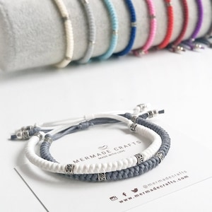 Könnte beinhalten: Zwei geflochtene Freundschaftsarmbänder, eines weiß und eines grau, mit silbernen Perlen. Die Armbänder befinden sich auf einer weißen Karte mit dem Text "Mermaid Crafts Made with Love" und den Social-Media-Handles @mermadecrafts und www.mermadecrafts.com.