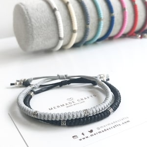 May include: Two friendship bracelets, one black and one grey, with silver beads and a knot at the end. The bracelets are on a white card with the text "Mermaid Crafts Made with Love" and social media information.