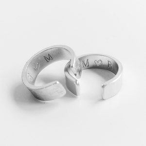 May include: Two silver rings with engraved initials and a heart symbol. The rings are interlocked, suggesting a couple's rings.