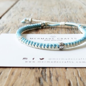 May include: A blue and white braided bracelet with silver beads. The bracelet is on a white card with the text "Mermaid Crafts Made with Love" and the website address "www.mermaidcrafts.com".