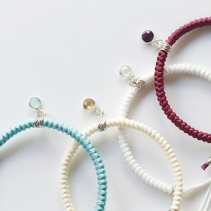 May include: Four braided bracelets with different coloured cords and gemstone charms. The bracelets are white, red, blue, and cream.