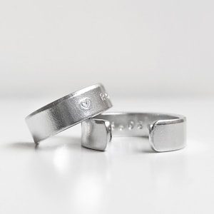 Op de afbeelding: Twee zilveren ringen met een gegraveerd hart op een van de ringen. De ringen zijn open en kunnen worden aangepast aan verschillende vingermaten.