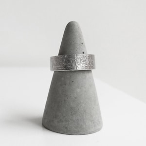 Può includere: Un anello d'argento con una superficie testurizzata, esposto su un porta anelli in cemento grigio.