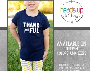 May include: A navy blue t-shirt with white text that says "THANK FUL" with a row of small white feathers underneath the word "FUL".