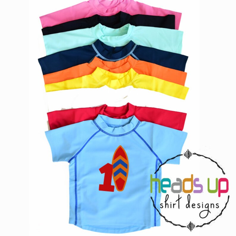 Op de afbeelding: Een stapel van tien kleurrijke rashguard shirts met een blauw shirt onderaan met een rood nummer &eacute;&eacute;n en een surfplank afbeelding. De tekst "heads up shirt designs" staat in de rechter benedenhoek van de afbeelding.
