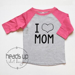 K&ouml;nnte beinhalten: Ein graues und pinkfarbenes Baseball-T-Shirt mit dem Text "I love MOM" in Schwarz. Das Herz ist schwarz umrandet.