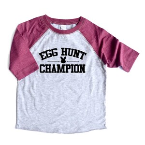 Peut inclure: Un t-shirt &agrave; manches raglan gris chin&eacute; et bordeaux avec le texte "Egg Hunt Champion" et un graphique de lapin.