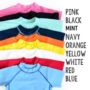 Op de afbeelding: Een stapel van acht rashguard shirts in verschillende kleuren, waaronder roze, zwart, mint, marine, oranje, geel, wit, rood en blauw. De shirts zijn gerangschikt van boven naar beneden in de volgorde van de vermelde kleuren. De tekst "PINK, BLACK, MINT, NAVY, ORANGE, YELLOW, WHITE, RED, BLUE" staat verticaal aan de rechterkant van de afbeelding.