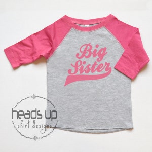 May include: A pink and grey long-sleeved shirt with a pink baseball-style graphic that says "Big Sister".