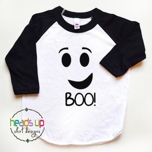 K&ouml;nnte beinhalten: Wei&szlig;es und schwarzes Baseball-Shirt mit einem Geistergesicht und dem Wort "BOO!" auf der Vorderseite.