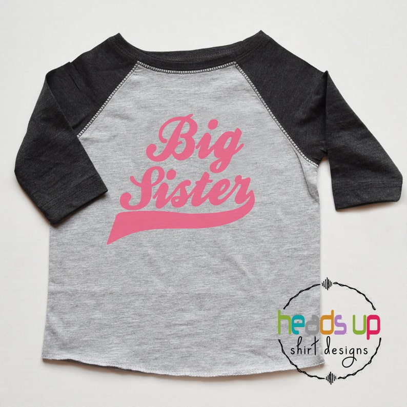 May include: A grey and black baseball tee shirt with pink lettering that says "Big Sister" in a retro font.