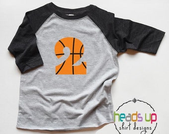 Può includere: Una maglietta da baseball grigia e nera con un numero "2" a forma di pallacanestro stampato sul davanti. La pallacanestro è arancione con linee nere.