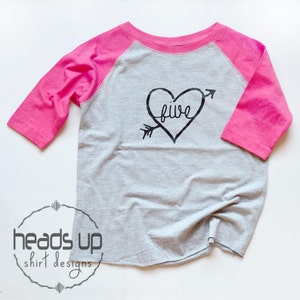 May include: A gray and pink baseball tee shirt with a black heart design that says "five" inside the heart. The heart has an arrow going through it.