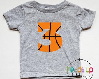 Può includere: Una maglietta grigia a maniche corte con un grande numero "3" a forma di palla da basket arancione sul davanti. La maglietta è fatta per un bambino.