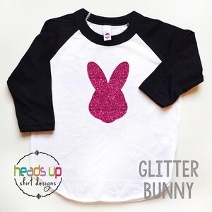 May include: A white and black baseball tee shirt with a pink glitter bunny silhouette on the front. The shirt has the text "GLITTER BUNNY" printed in gray below the bunny. The text "heads up shirt designs" is printed in a circle on the bottom left of the shirt.
